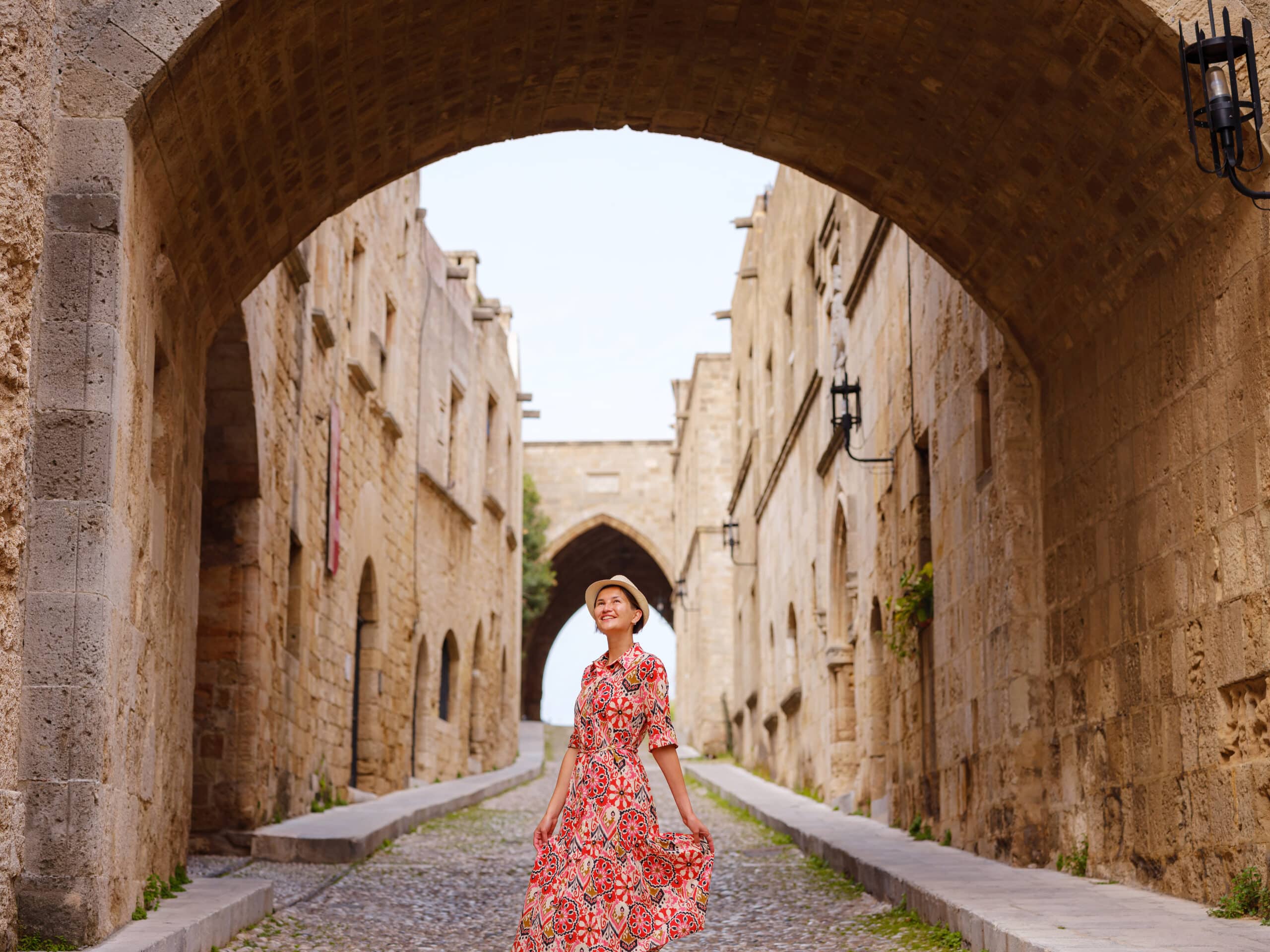 summer trip to Rhodes island, Greece. Young Asian woman in ethnic red dress walks Street of Knights of Fortifications castle. female traveler visiting southern Europe. Unesco world heritage site.